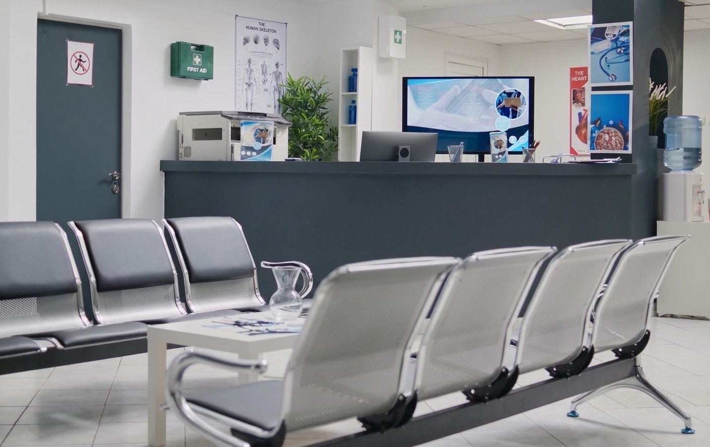 Salle d'attente d'un cabinet de médecine générale : rangées de sièges vides devant un comptoir d'accueil.