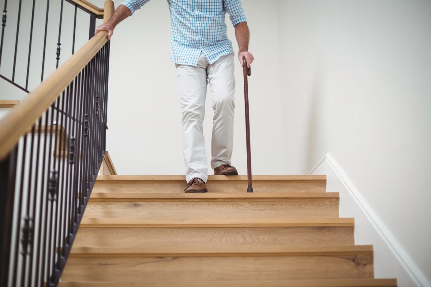 Une personne âgée descend un escalier en bois en se tenant à la rampe, une canne à la main.