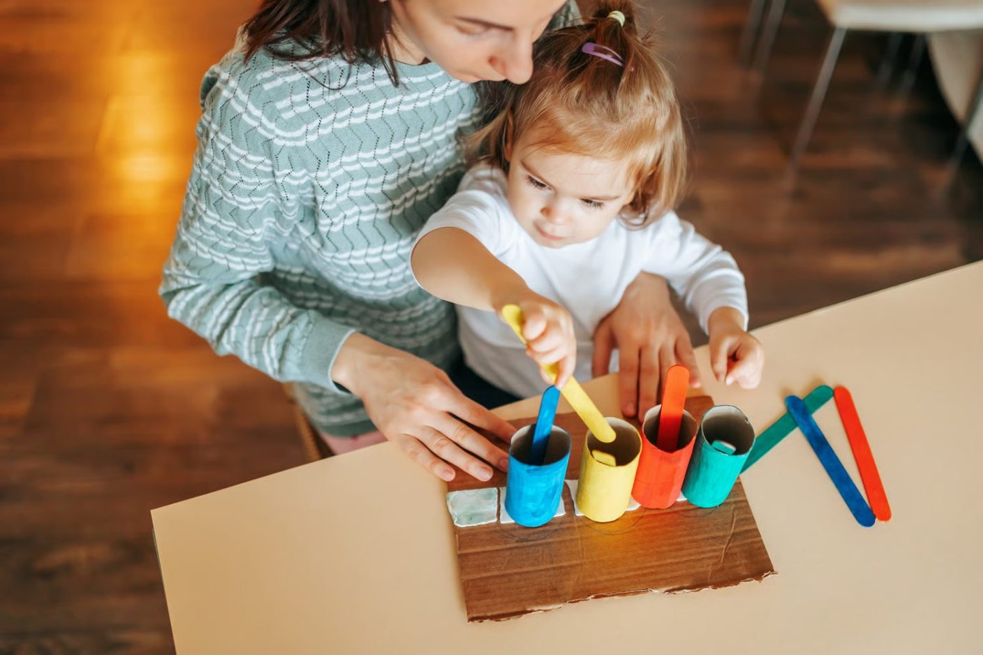 Une petite fille assise sur les genoux d'une adulte glisse des bâtonnets colorés dans des rouleaux de la même couleur.