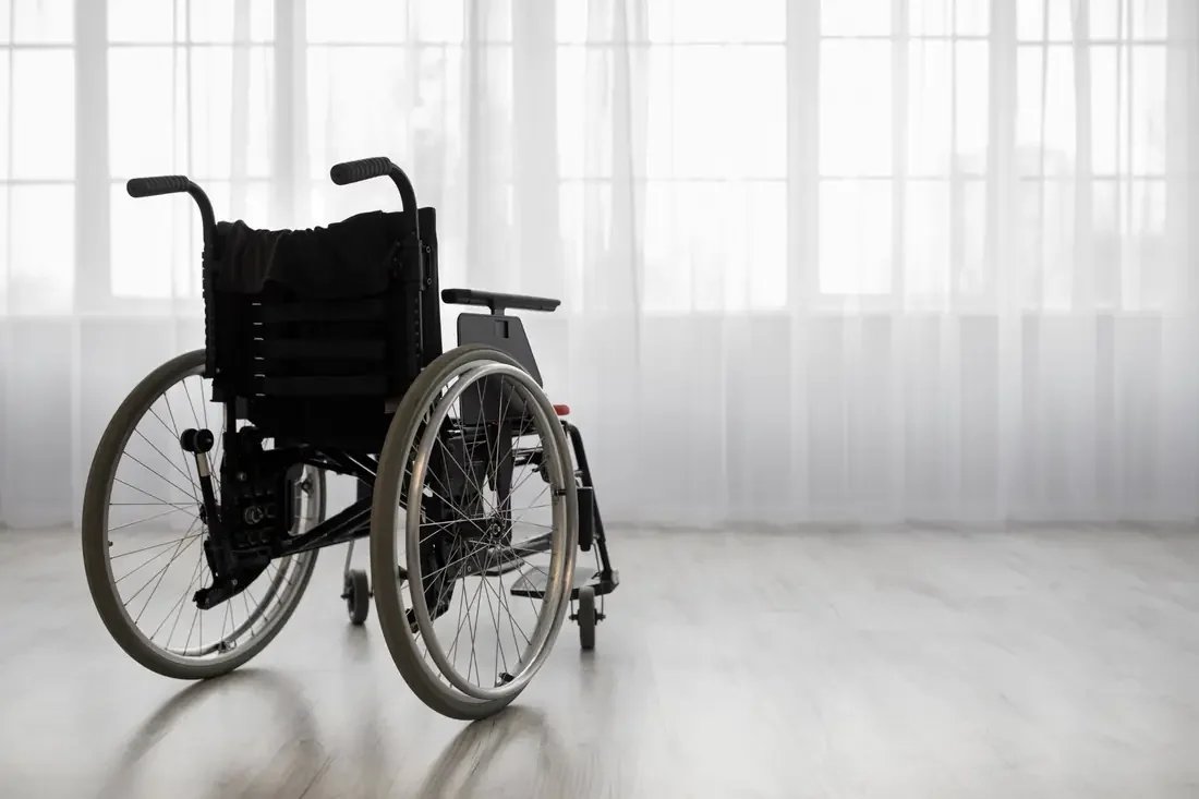 Fauteuil roulant manuel vide devant une baie vitrée voilée de rideaux blancs.